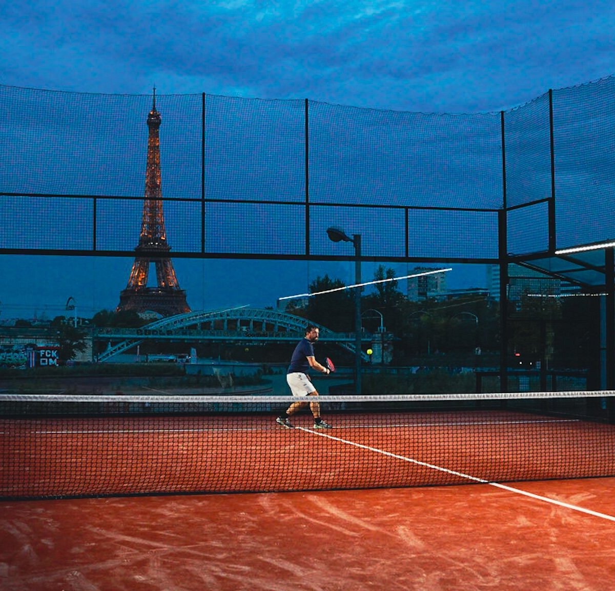 Un joueur sur un court en terre battue au bord de la Seine, à la tombée de la nuit, la tour Eiffel illuminée au fond.